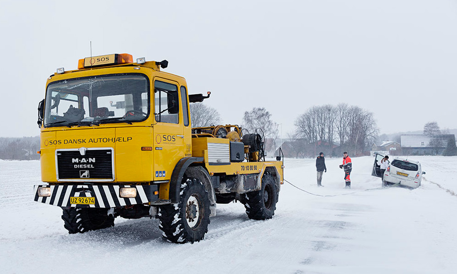 Redderbil og to personer arbejder ved en bil, der er kørt i grøften under snevejrforsøg.